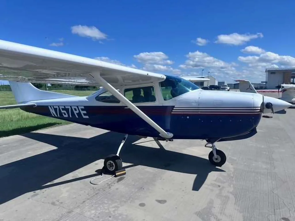 A white and blue Cessna 172, tail number N757PE, parked on an airfield under a blue sky.