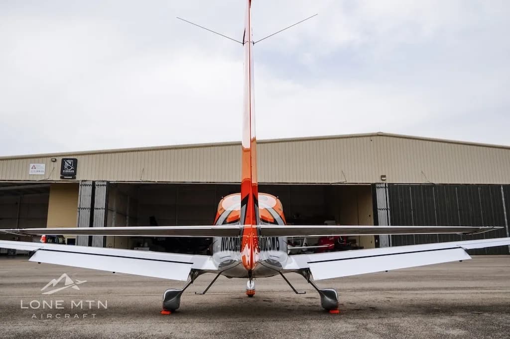A rear view of the orange and silver Cirrus SR20 aircraft N685MB parked in front of a hangar.
