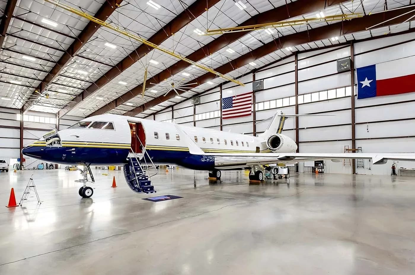 A large business jet with a blue and gold stripe livery, parked inside a hangar with its door open and stairs extended.