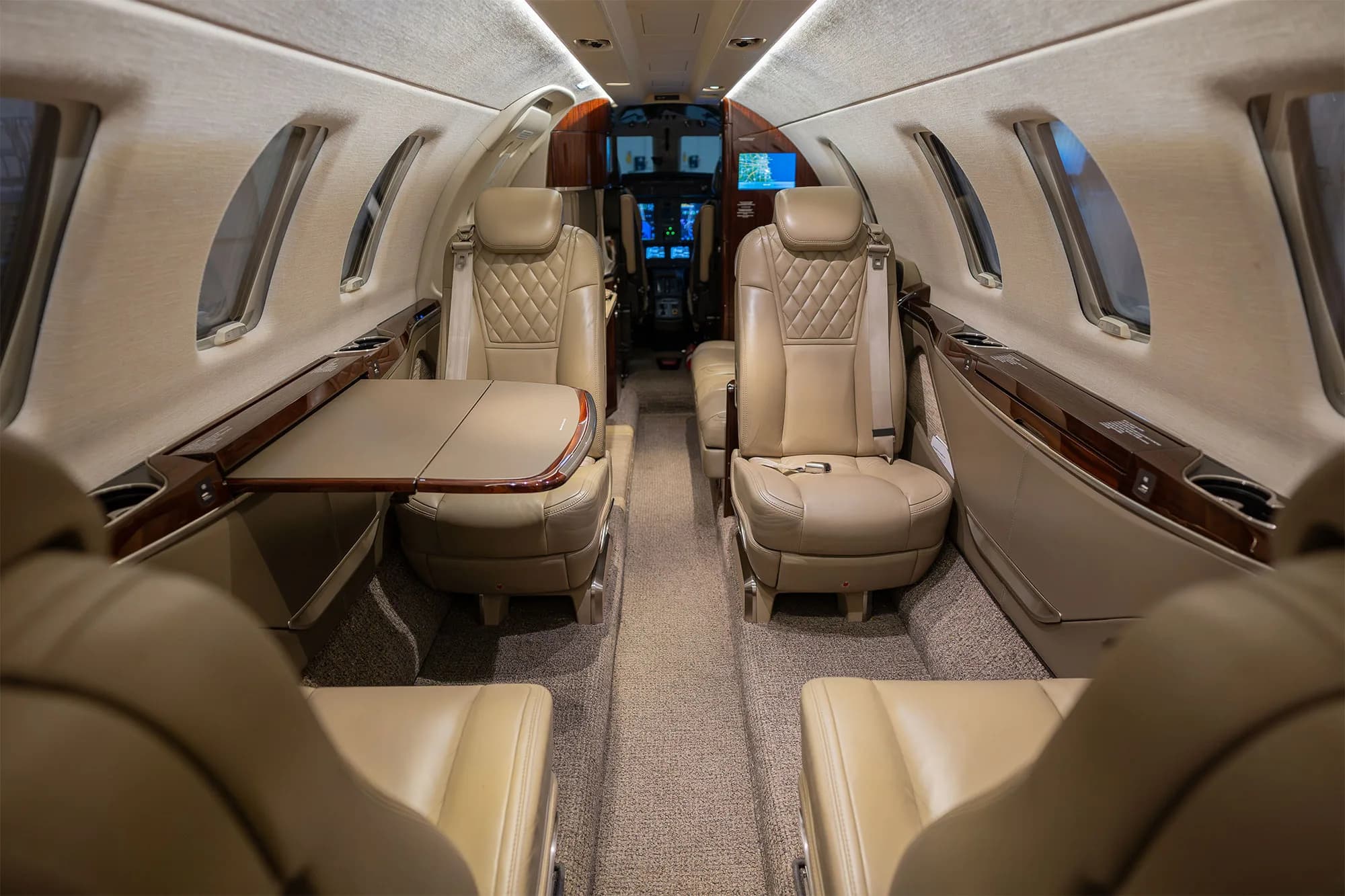 The interior cabin of a business jet with beige leather seats and a deployed side table, looking towards the cockpit.