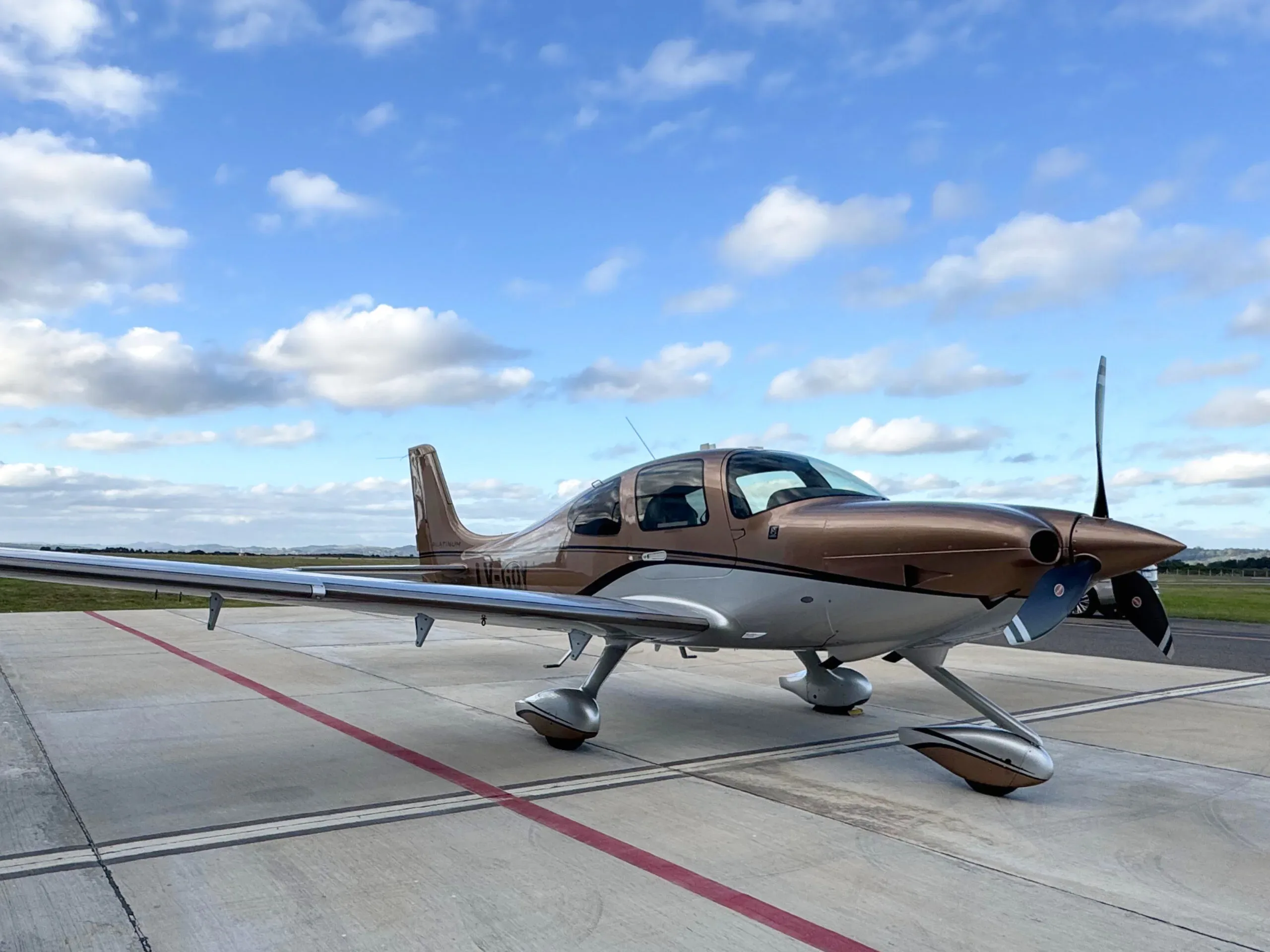 A brown and silver Cirrus SR22T aircraft parked on an airport tarmac under a cloudy sky.
