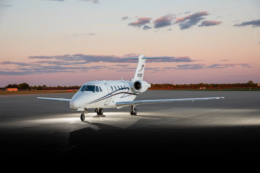 A white Cessna Citation business jet with blue and red stripes on the tail, viewed from the front-right on a tarmac at sunset.