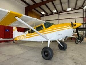 A yellow and white Cessna 185 Skywagon, registration N185GS, is parked inside a hangar.