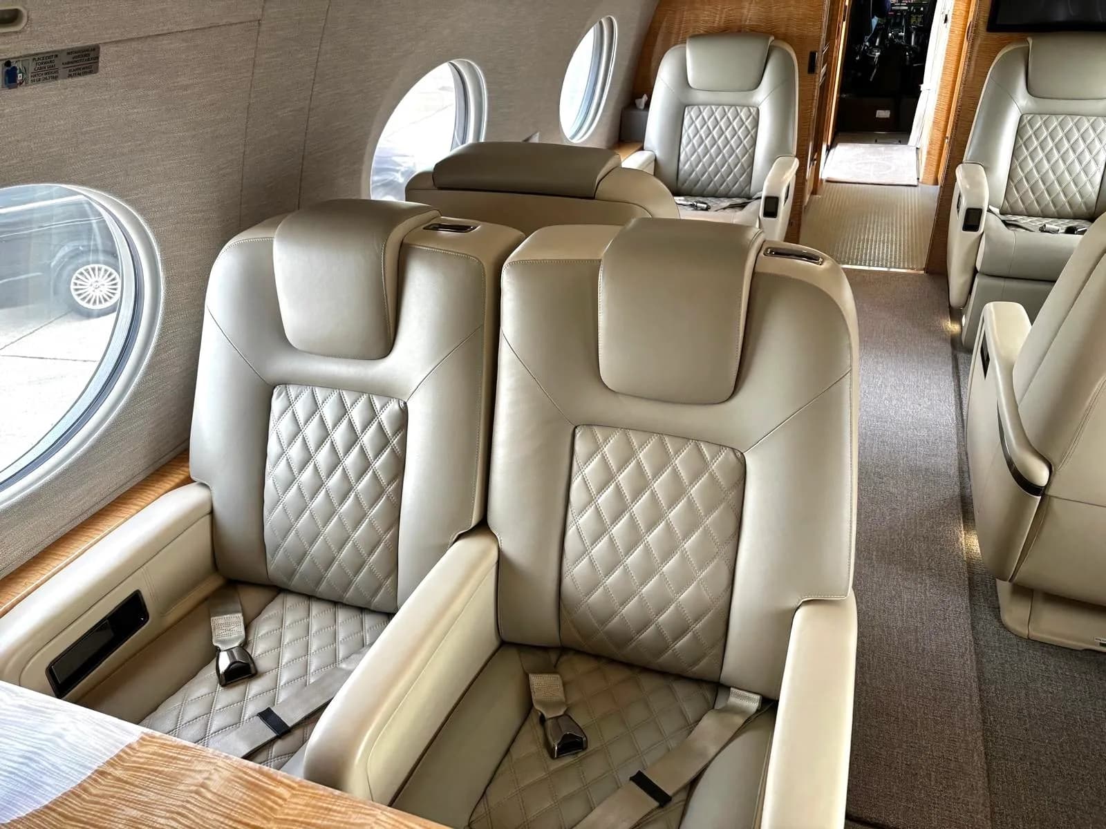 A close-up view of two light-colored quilted leather seats inside a private jet cabin, with a window visible.