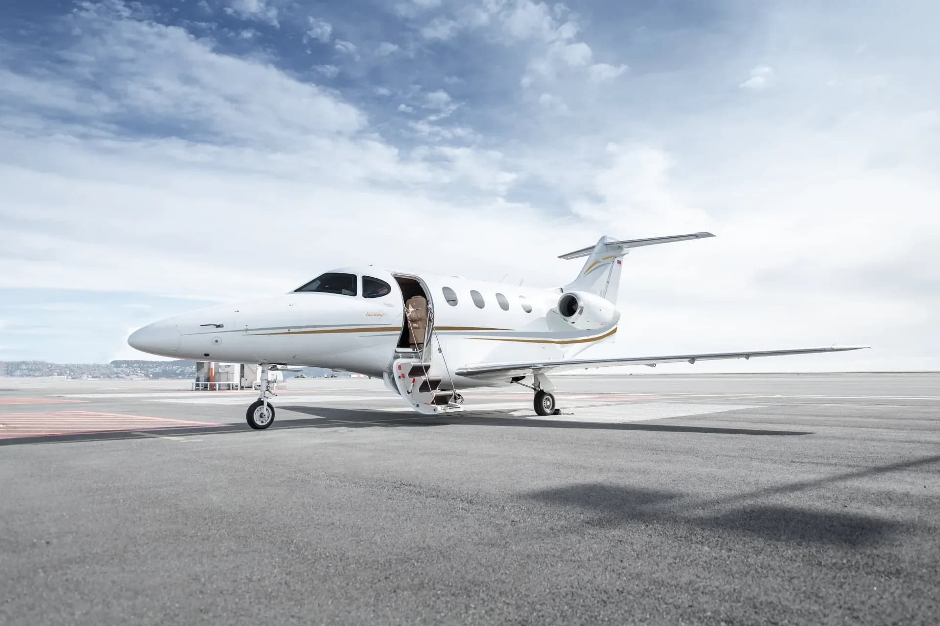 A white business jet with gold accents and its main door open, parked on an airport tarmac under a partly cloudy sky.