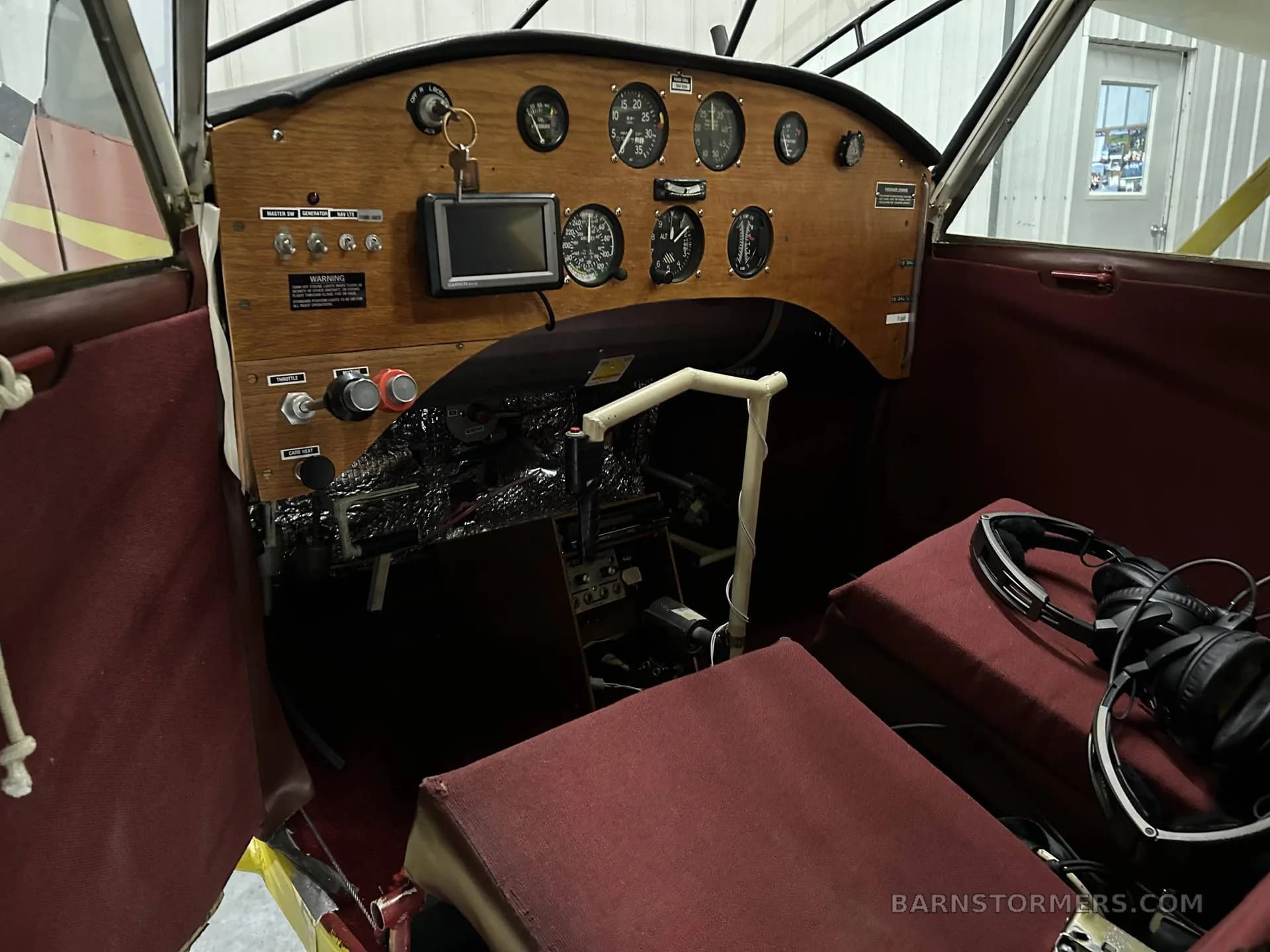 The cockpit of an aircraft features a wooden instrument panel, a yoke, and red fabric seats with headphones.