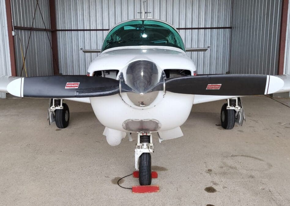A head-on view of a white Mooney M20J aircraft with a black propeller, parked inside a hangar.