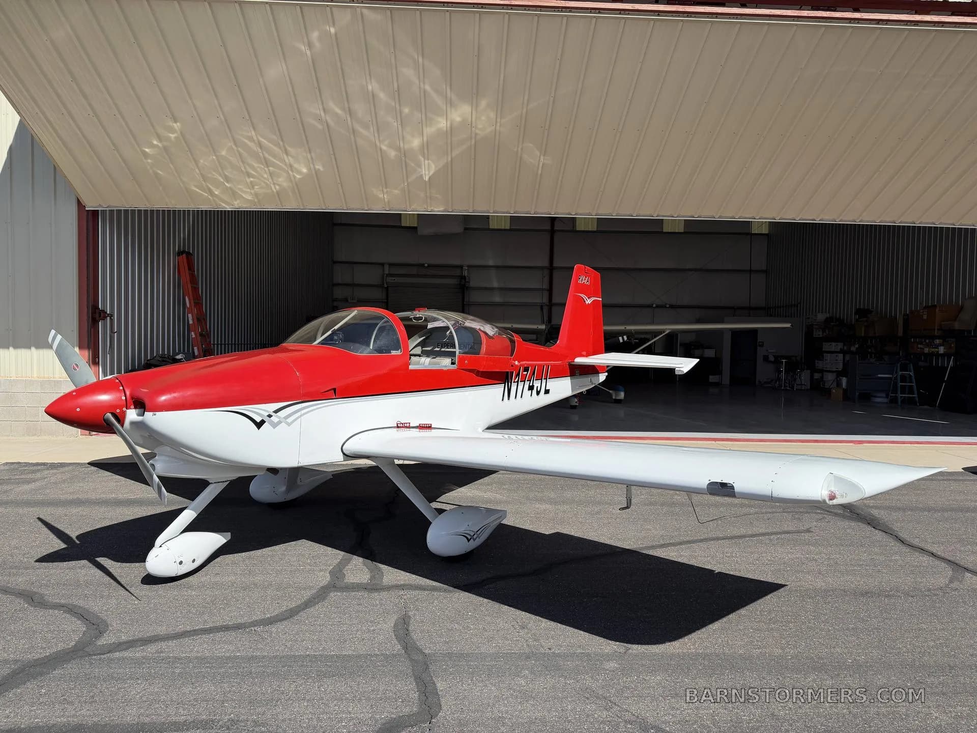 A red and white Vans RV-7A aircraft, registration N174JL, is parked on the tarmac in front of an open hangar, viewed from the side.