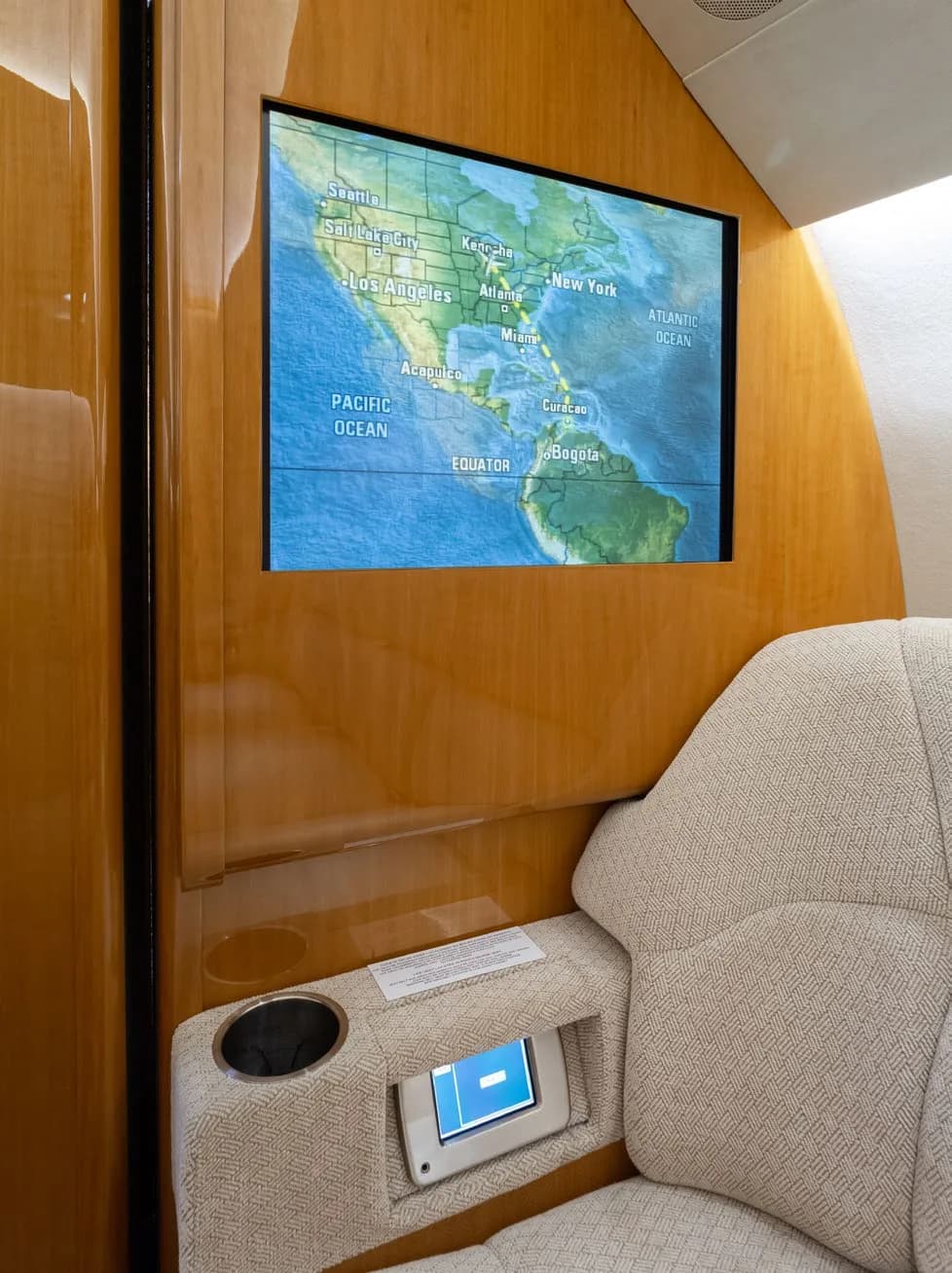 A cabin screen displaying a world map with flight path information, mounted on a polished wooden wall next to a textured beige seat.