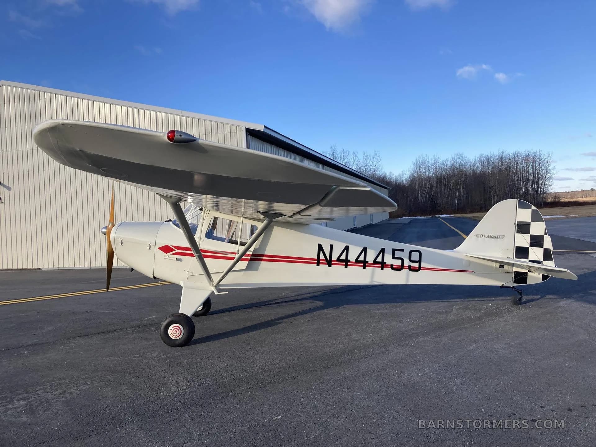 A white and red Piper Cub N44459 with a checkered tail, parked on an asphalt apron next to a hangar under a blue sky.