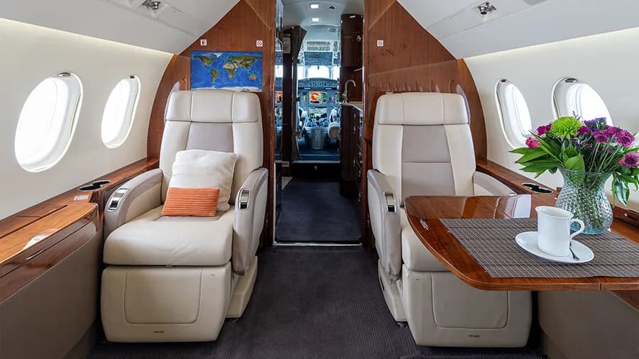 The forward cabin of a private jet features two beige leather seats, a small table with flowers and a teacup, and dark wood accents.