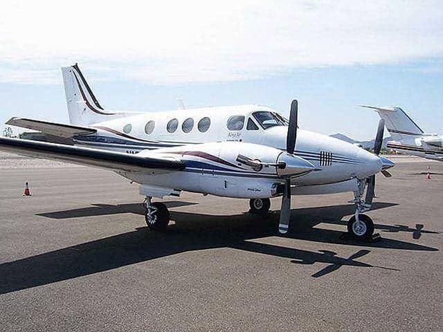 A white Beechcraft King Air B200 with blue and maroon stripes is parked on an airfield with another aircraft in the background.