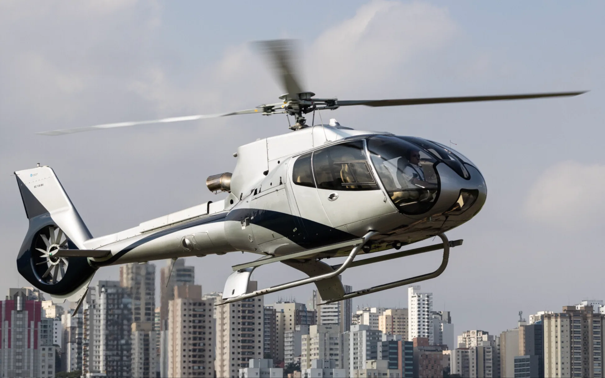 A silver and black Eurocopter EC130 B4 helicopter in flight over a city.
