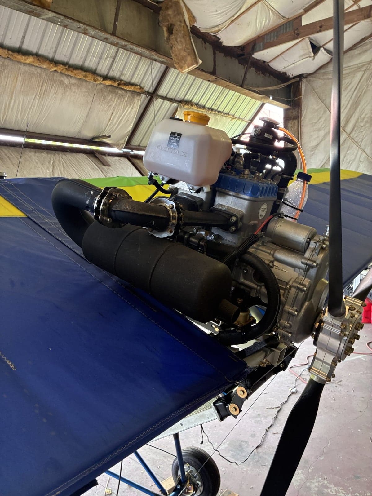 Close-up view of the Rotax engine and propeller mounted on the wing of a blue and yellow ultralight aircraft in a hangar.