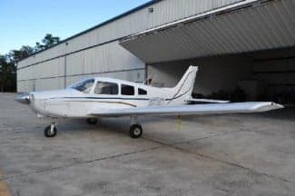 Side view of a white and silver Piper PA-28-181 Archer II aircraft, N6789T, parked in front of a hangar.