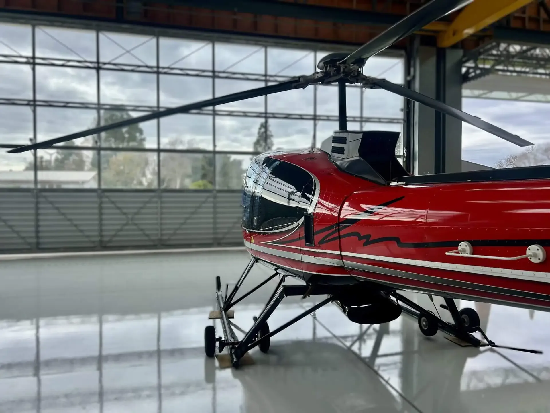 A red and black helicopter is positioned inside a hangar, resting on small ground handling wheels.