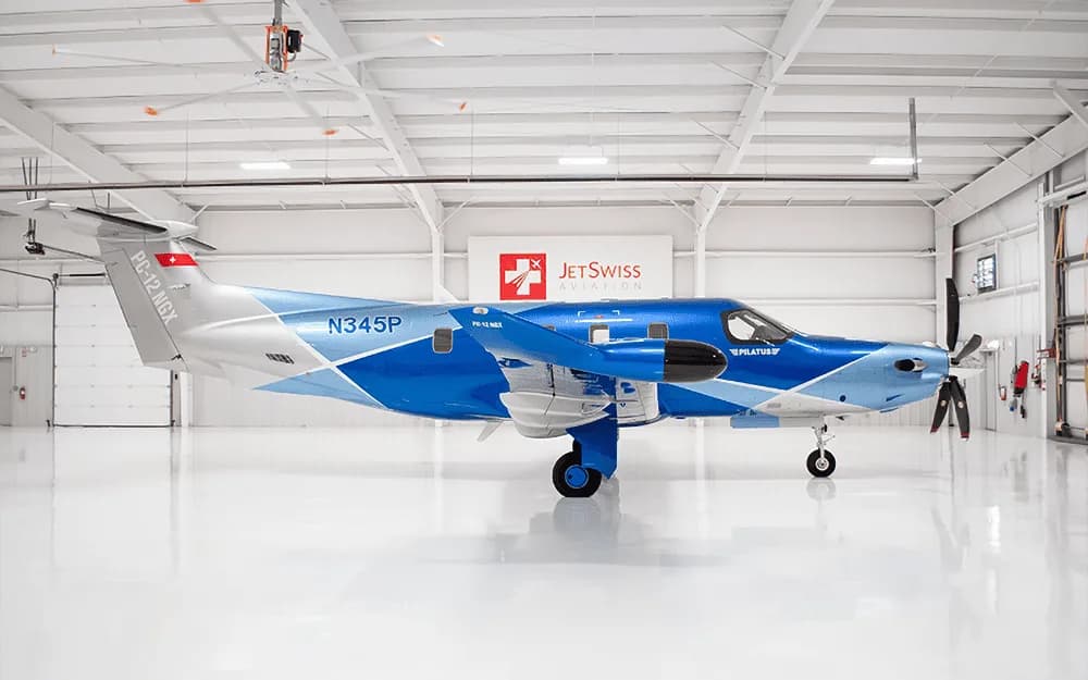A side view of a Pilatus PC-12 NGX aircraft with a blue and silver paint scheme, registration N345P, parked inside a white hangar.