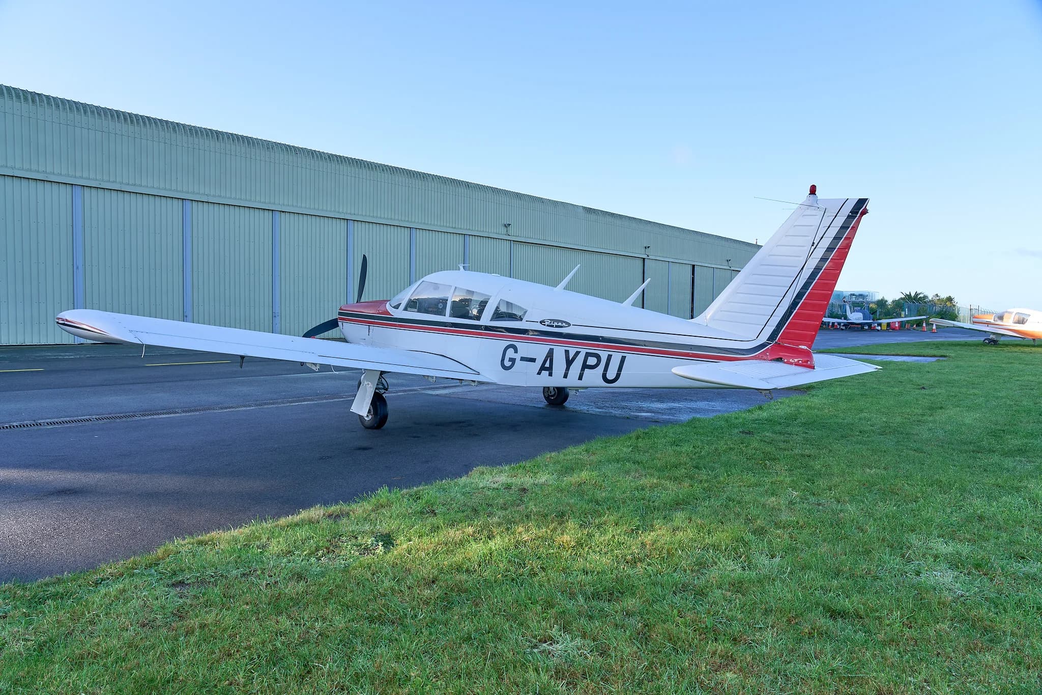 A side view of the white and red Piper Cherokee G-AYPU with a large green hangar in the background.