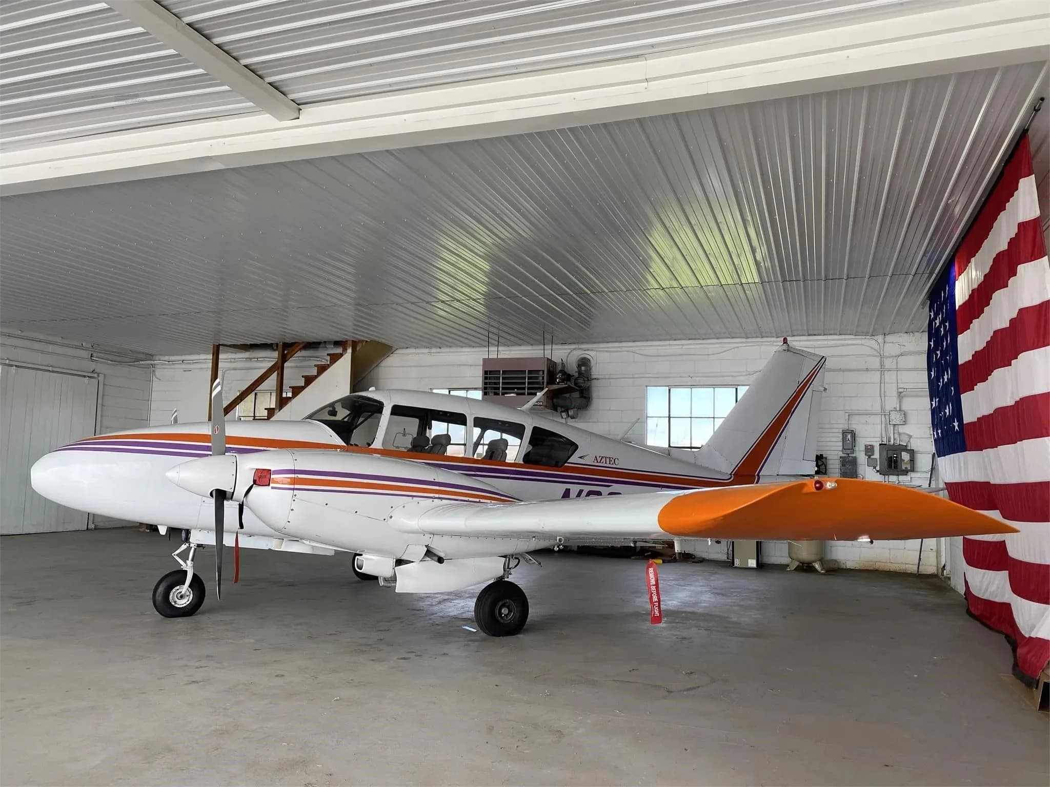 A side view of a white Piper Aztec twin-engine aircraft with orange and purple stripes in a hangar, showing the tail and right wing.
