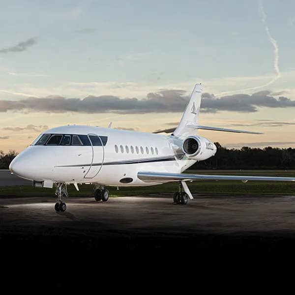 A white Dassault Falcon business jet with a dark stripe on the fuselage sits on a tarmac at sunset.