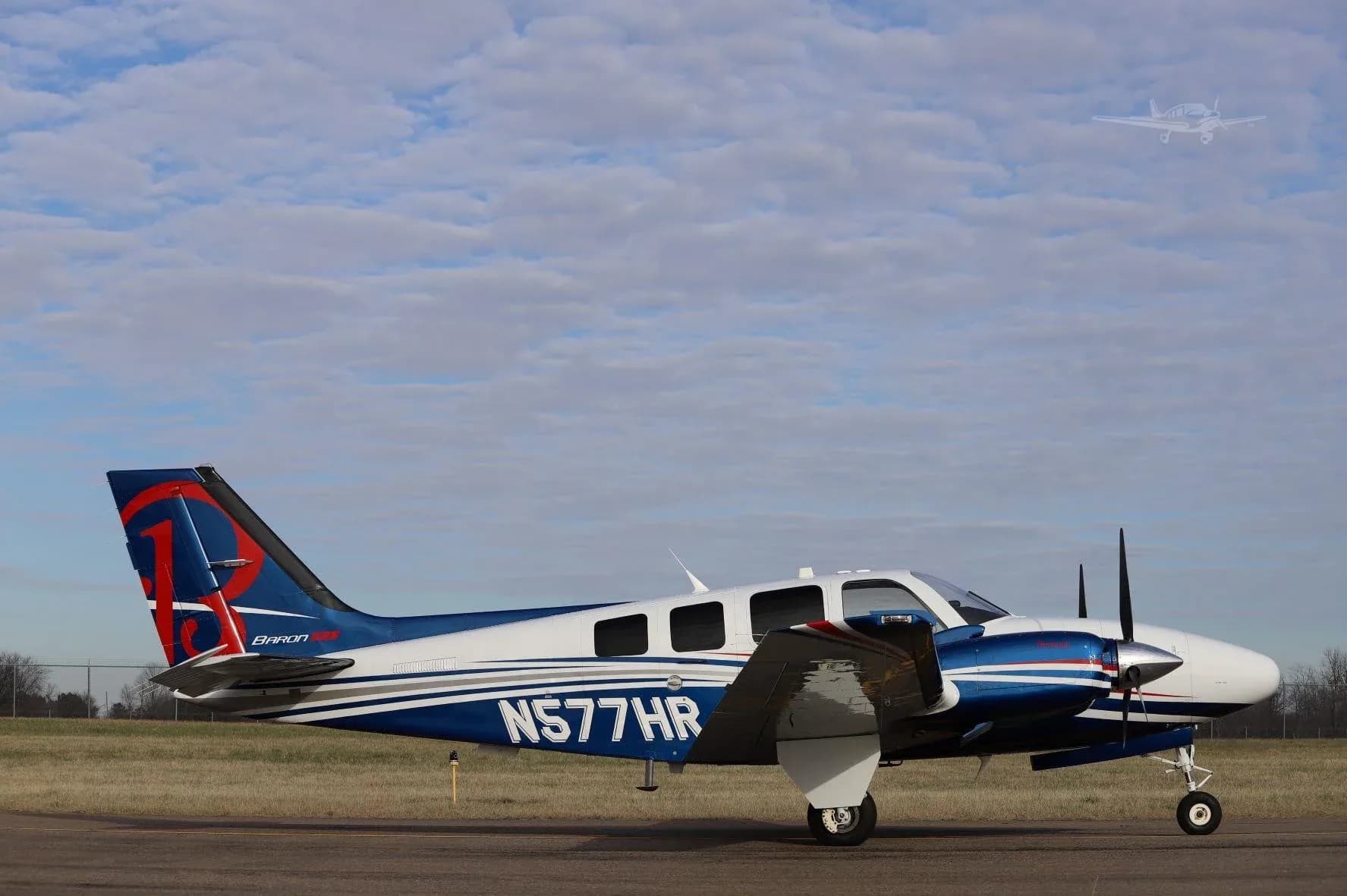 A white and blue Beechcraft Baron G58 (N577HR) with red tail accents, seen from the right side on a tarmac under a partly cloudy sky.