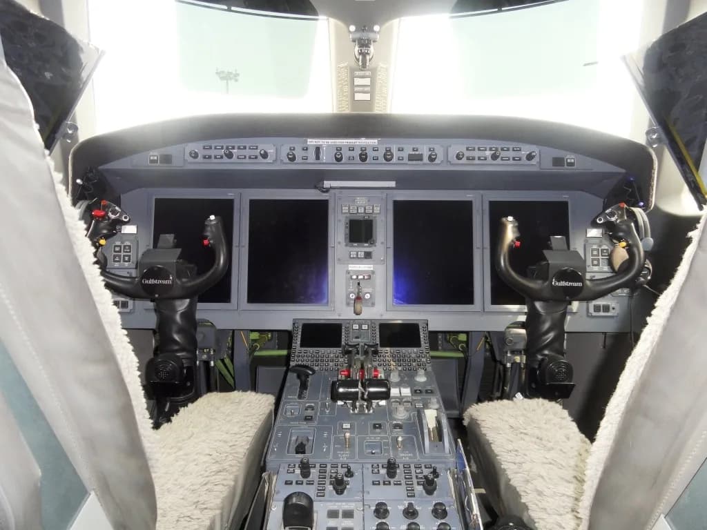 The cockpit of a Gulfstream aircraft, showing the instrument panel with multiple screens, yokes, and central console.