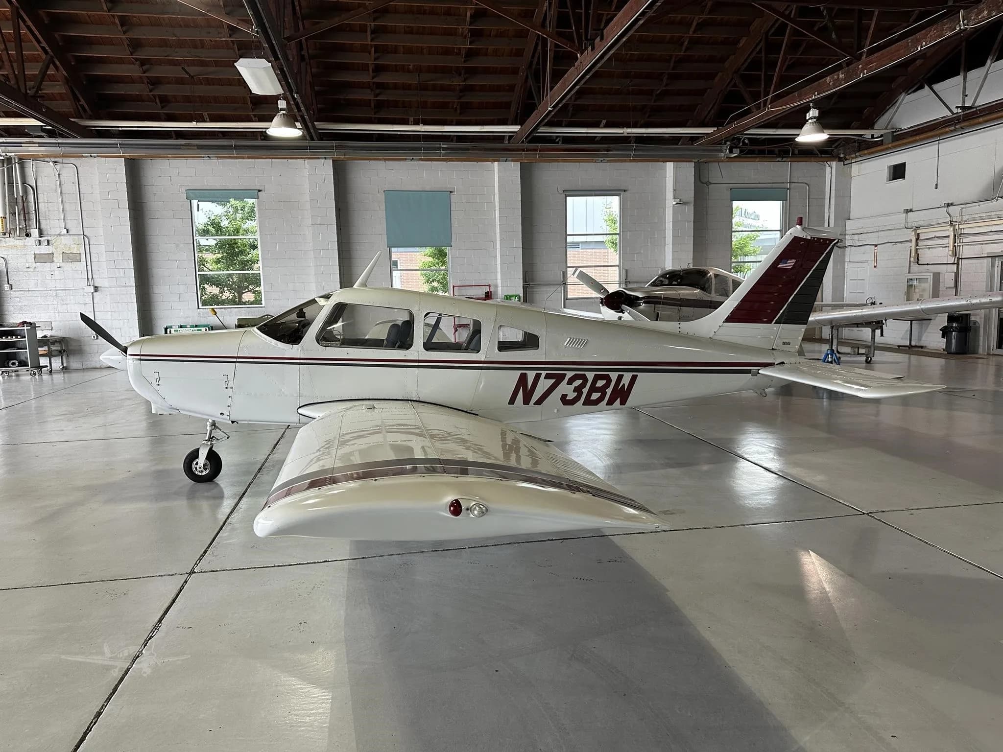 A white single-engine aircraft with maroon and black stripes, registration N73BW, viewed from the left side in a hangar.