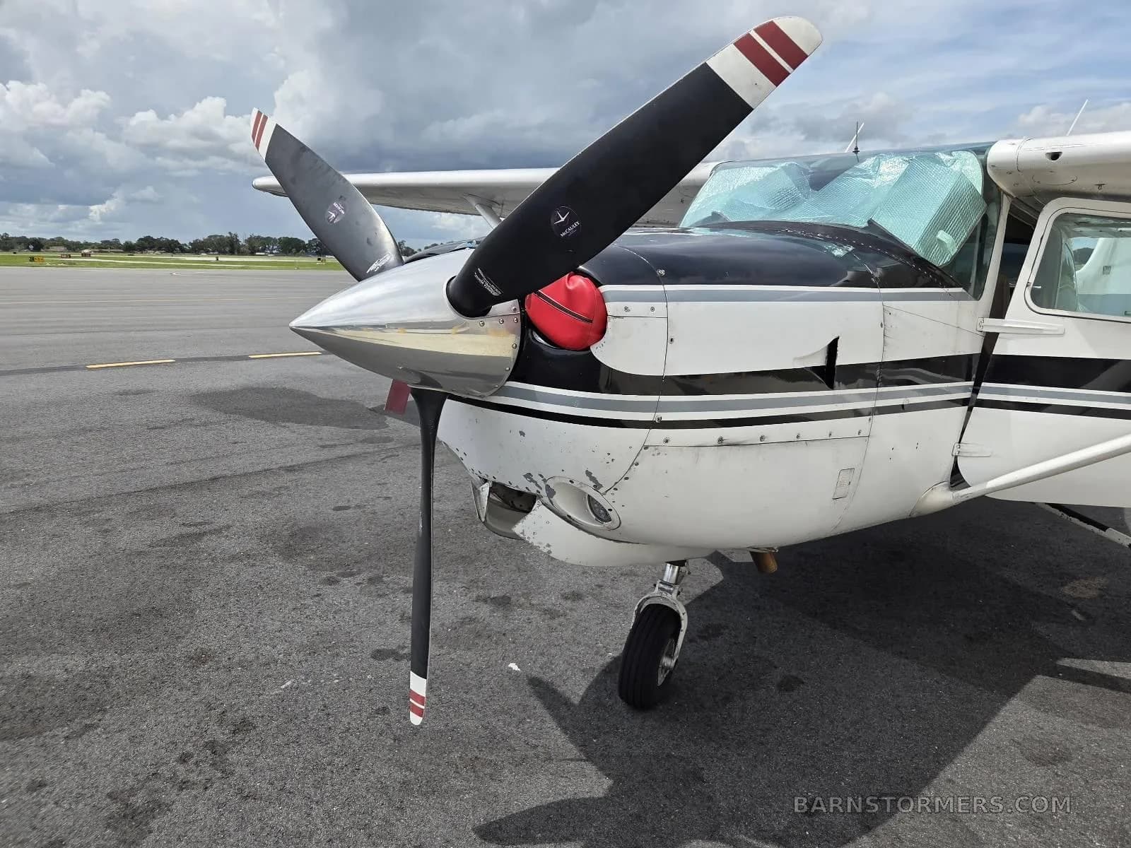 A front-quarter view of a white and black Cessna aircraft, highlighting the polished propeller, red spinner cover, and a sunshade in the windshield.