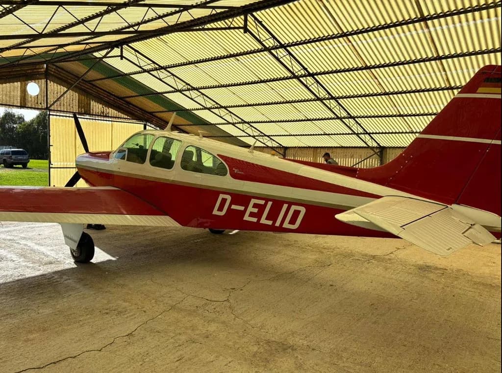 A side view of the red and white Beechcraft Bonanza D-ELID parked in a hangar, with a vehicle visible outside.