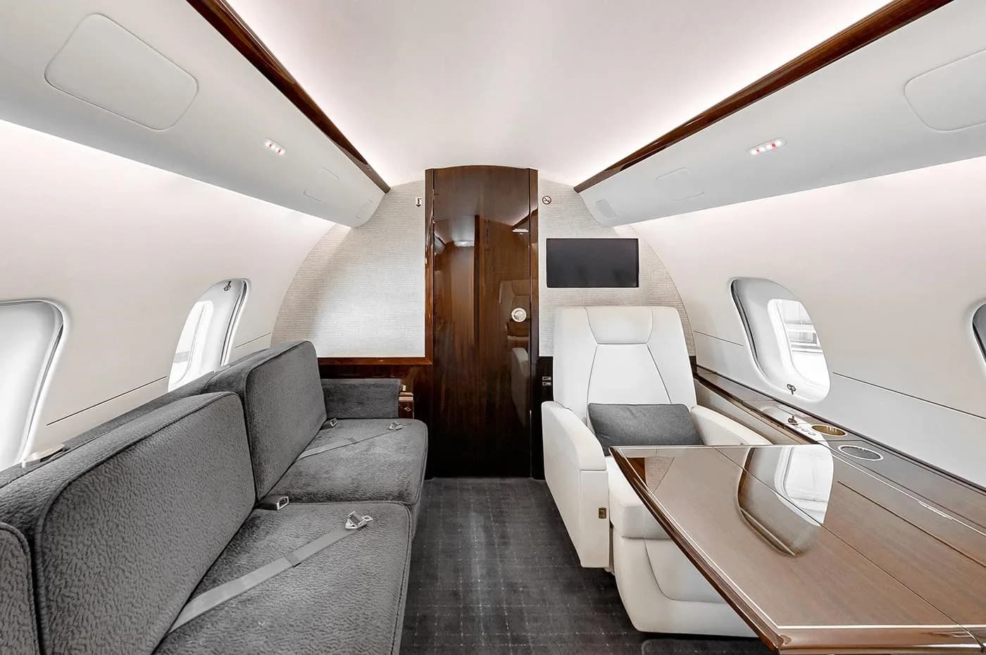 The interior cabin of a business jet, showing a grey sofa, a cream armchair, and a dark wood table with a mounted screen.