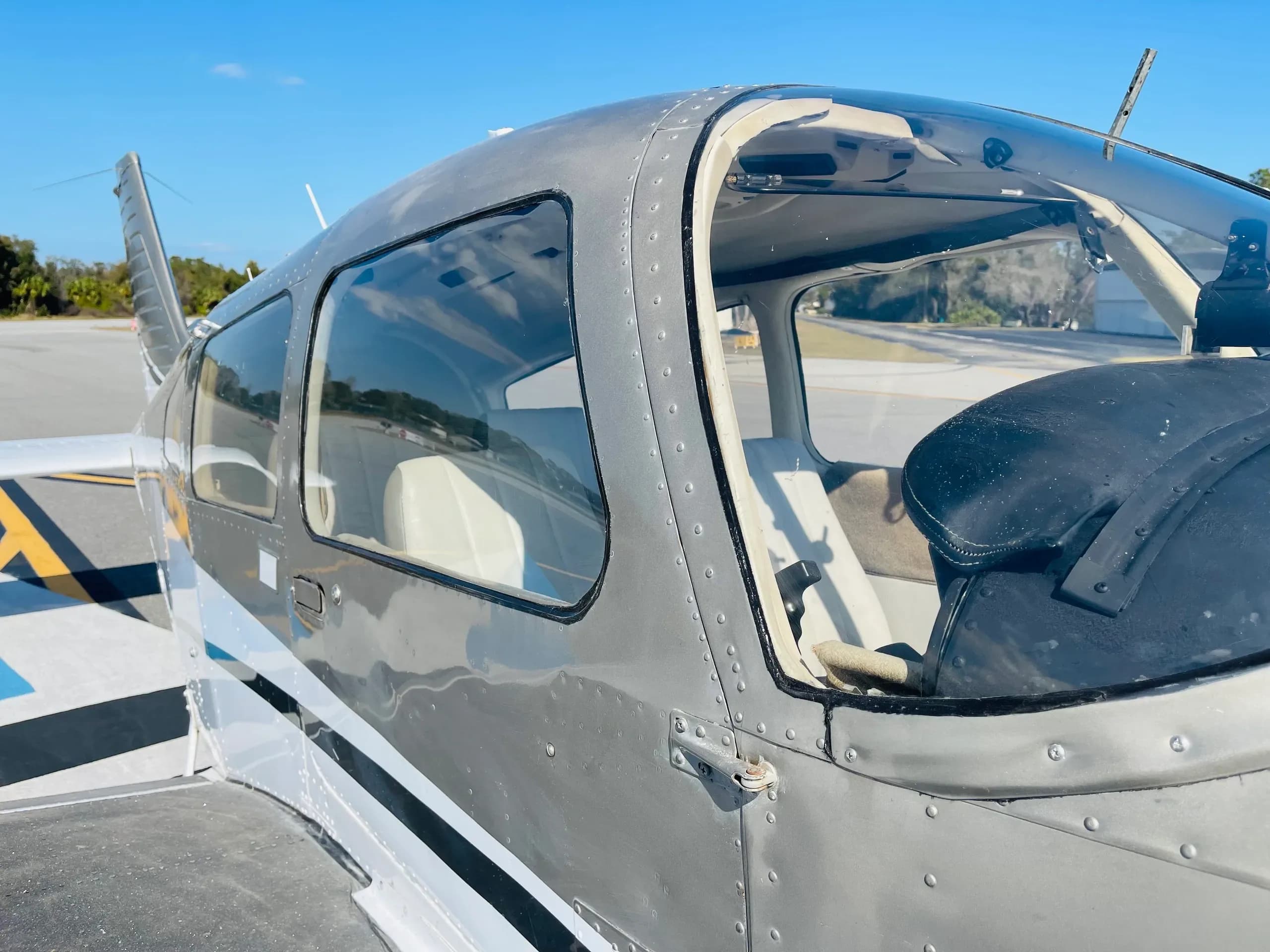 A close-up exterior view of the cockpit and cabin windows of a gray and white single-engine aircraft on an airfield.
