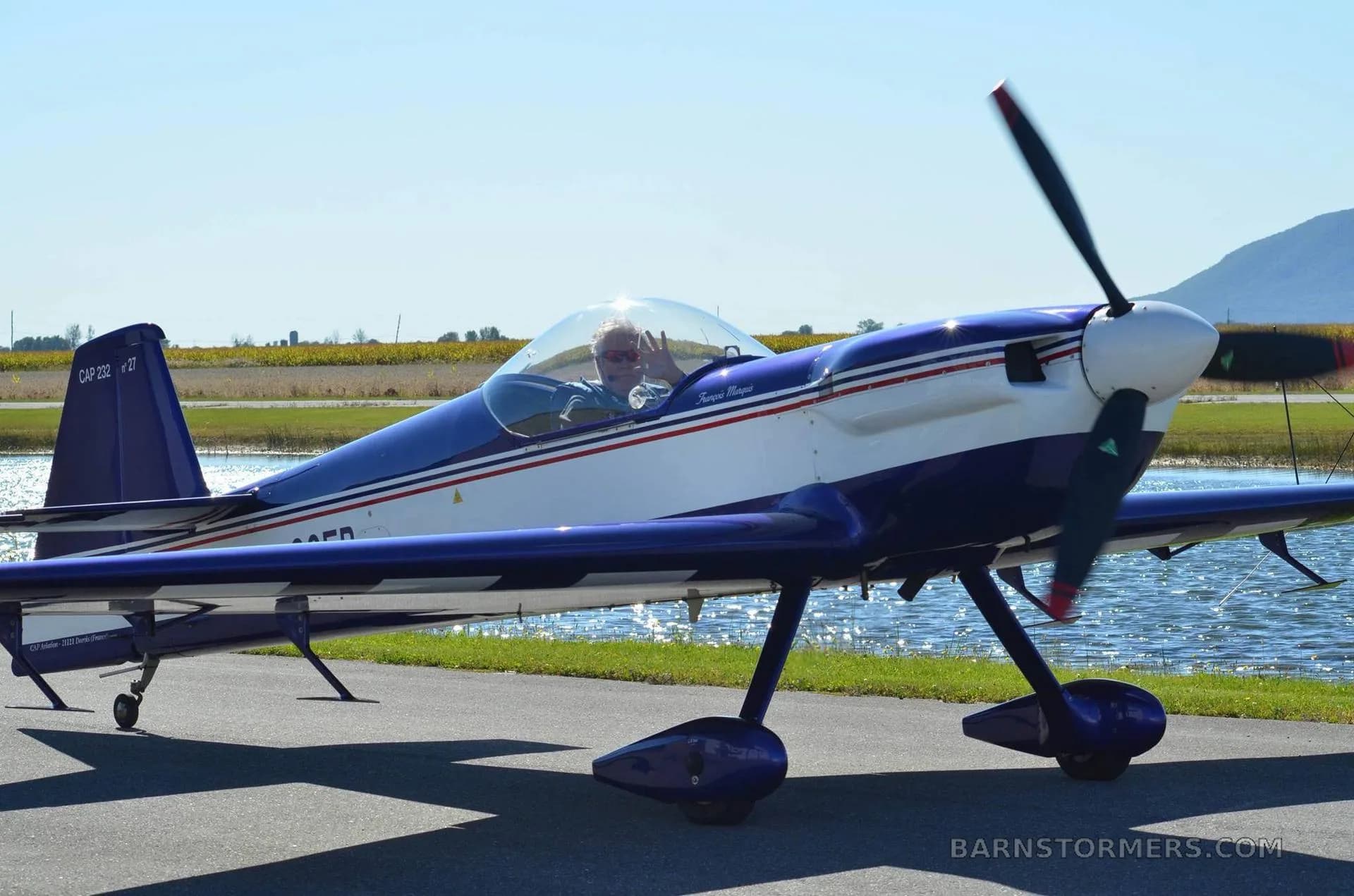 A blue and white CAP 10B aerobatic aircraft is on the ground next to water, with a pilot waving from the open canopy.