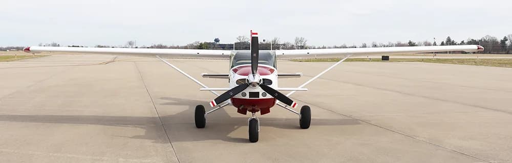 A front view of a white and red single-engine high-wing aircraft with a propeller, parked on an airfield.
