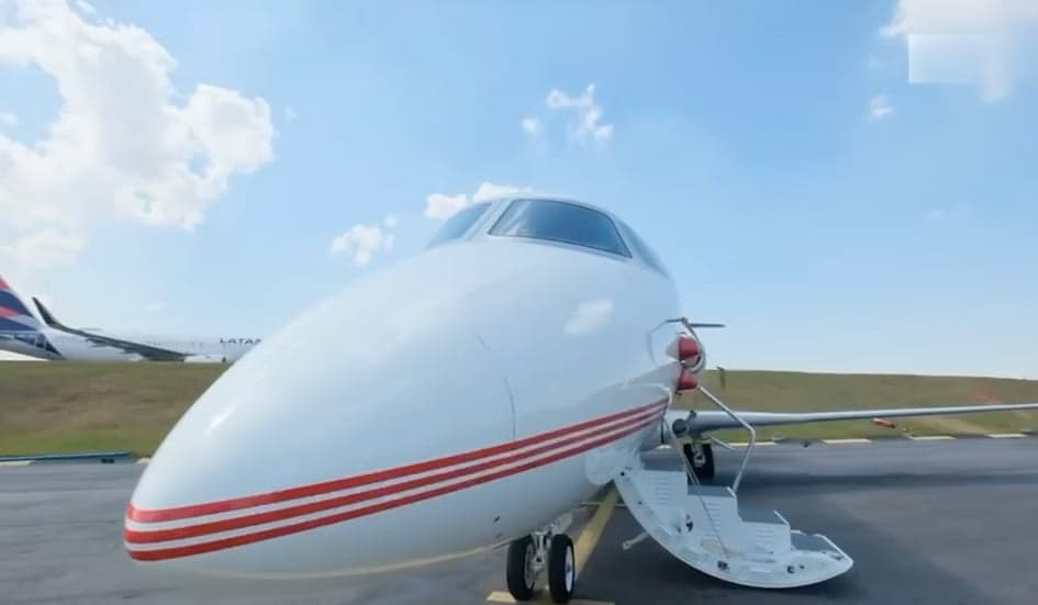 The front of a white business jet with red stripes, parked on an tarmac with its main door open, with a larger airliner in the background.