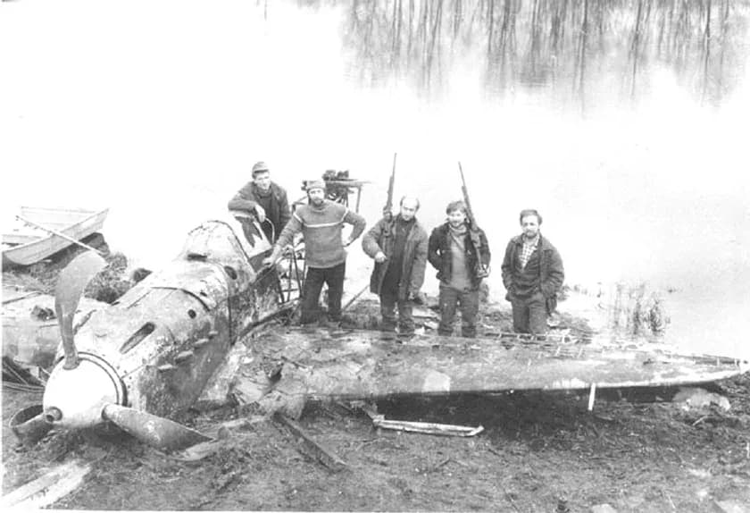 A group of people stand next to a crashed aircraft near a body of water.