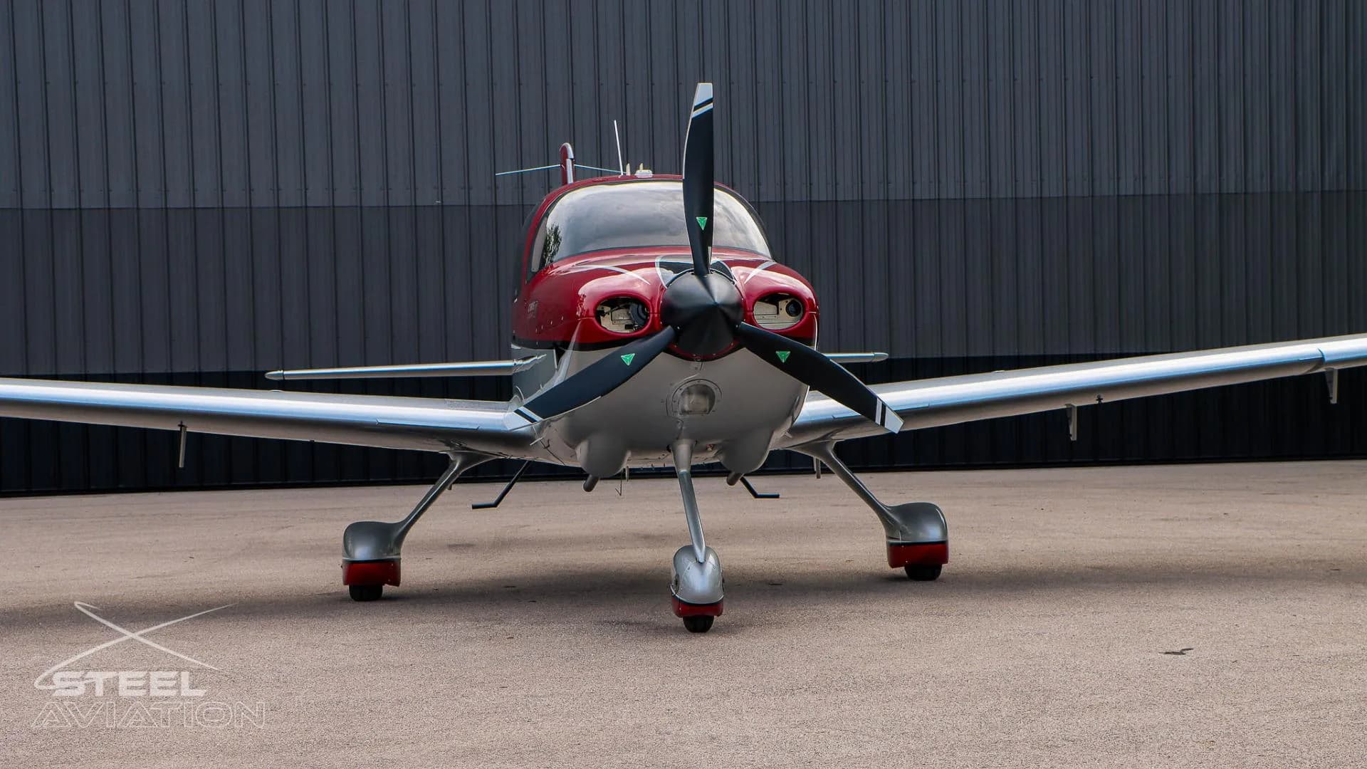 A close-up front view of the red and white Cirrus SR22 aircraft, showing its propeller and nose, parked in front of a dark gray hangar.
