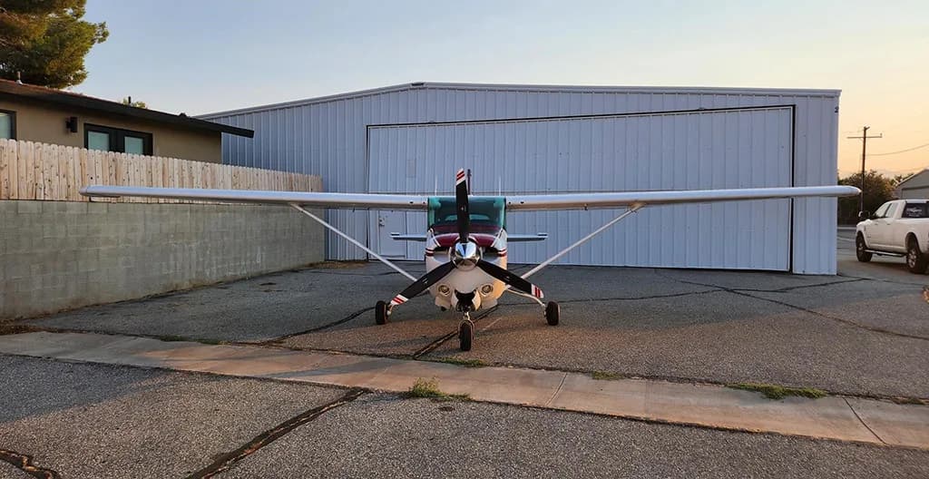 A front view of a white and maroon Cessna 172 aircraft parked on an asphalt apron in front of a hangar.