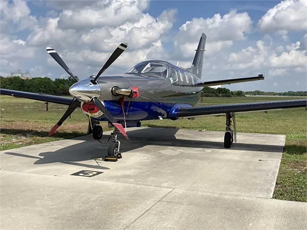 A grey and blue single-engine turboprop aircraft parked on a concrete pad under a partly cloudy sky.