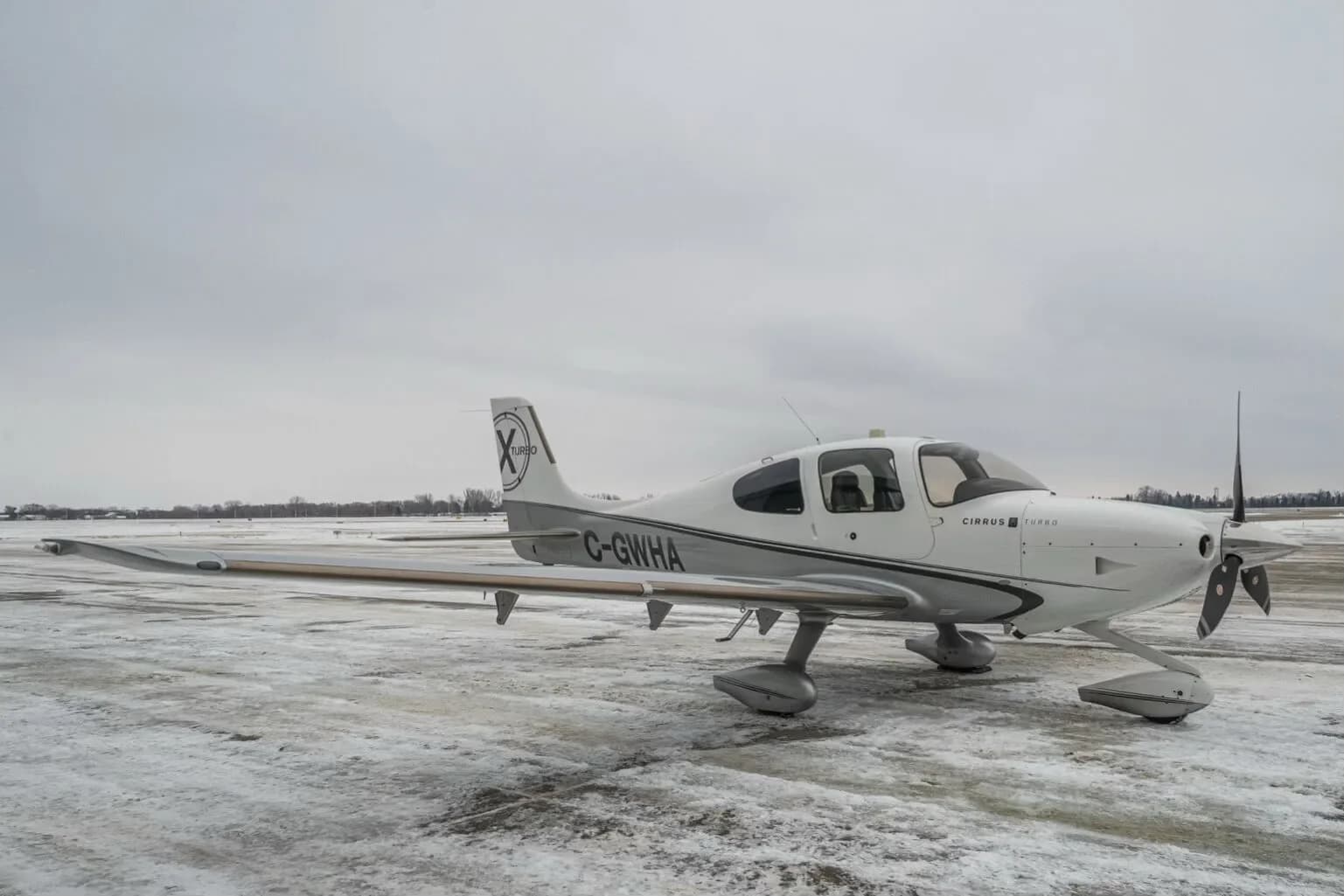 A white and grey Cirrus SR22T aircraft (C-GWHA) with a 'Turbo X' logo on the tail, parked on a snowy tarmac under an overcast sky.