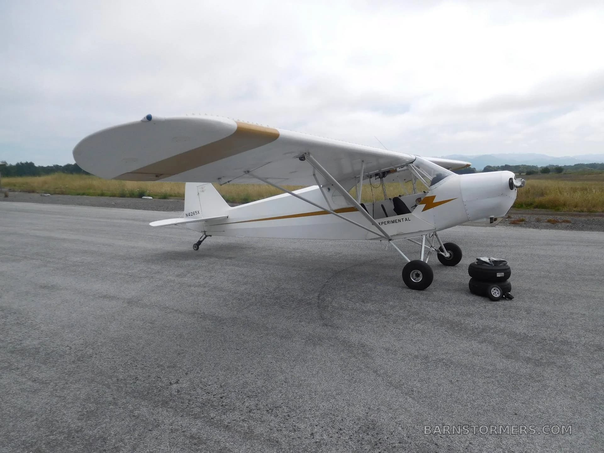 A white experimental aircraft with gold wing stripes is parked on an asphalt runway under a cloudy sky.