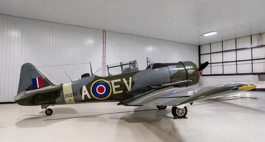 A North American Harvard Mk.IIB aircraft in green and grey camouflage with RAF roundels, parked in a hangar.