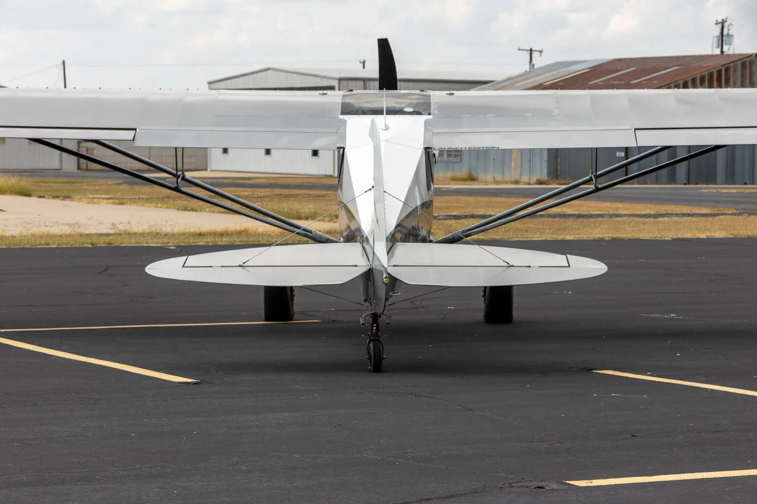 A rear view of the white X-Cub aircraft with its tailwheel, parked on an asphalt runway with hangars in the background.