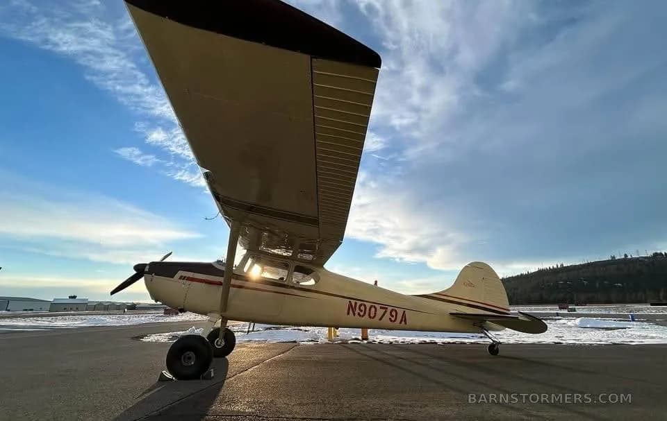 A cream and brown taildragger aircraft, N9079A, parked on a snowy tarmac with the sun low in the sky.