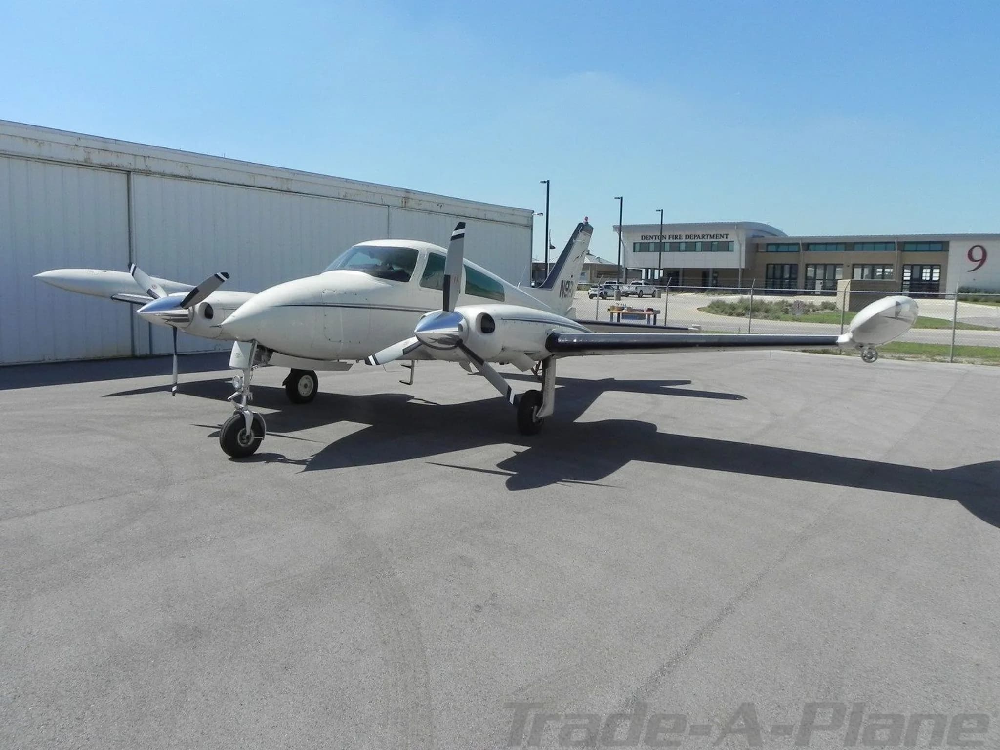 A white twin-engine Cessna 310 aircraft is parked on an asphalt apron with hangars and a fire department building in the background.