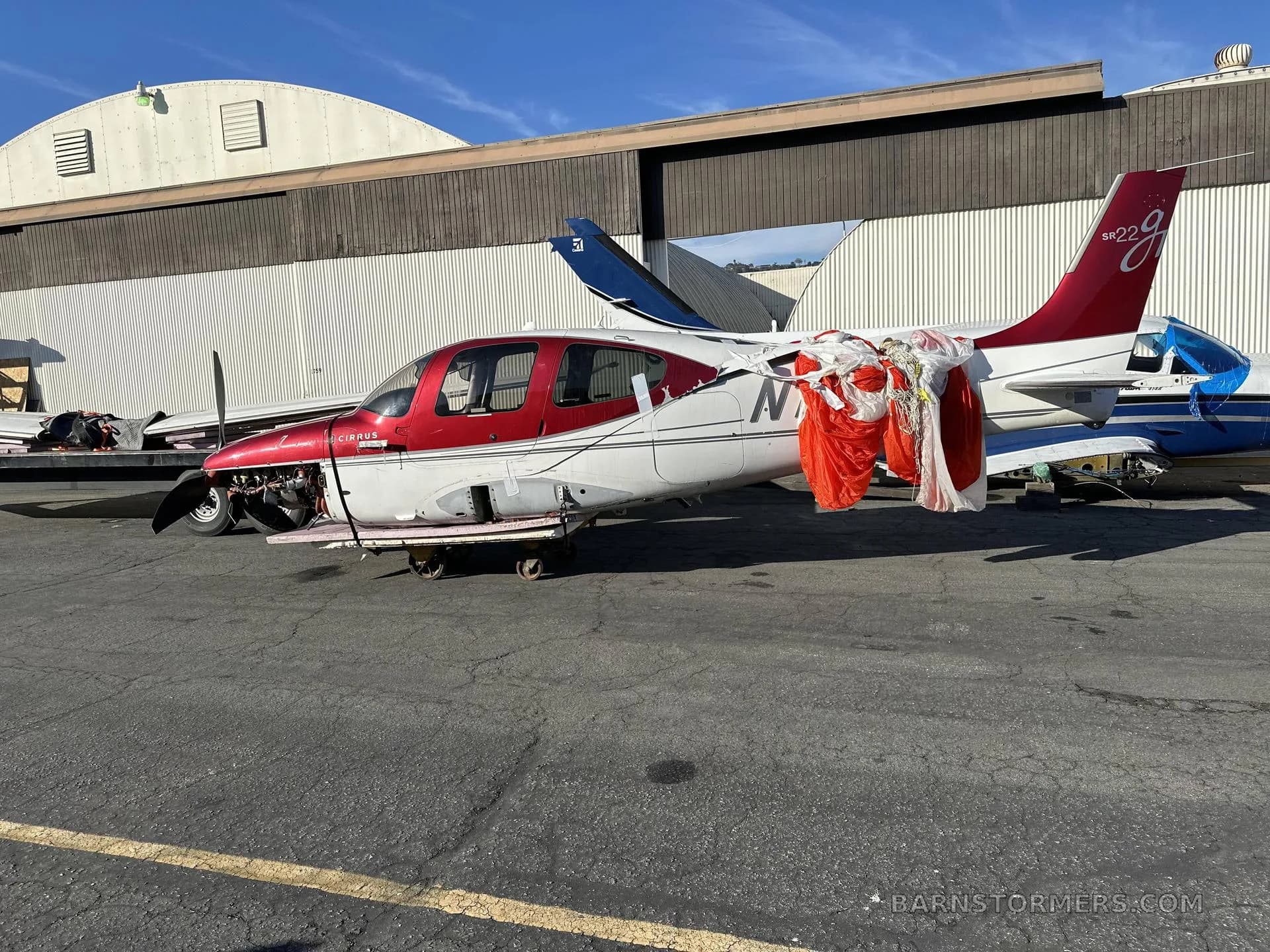 A red and white Cirrus SR22 aircraft with a deployed parachute draped over its fuselage, parked outdoors next to a hangar.