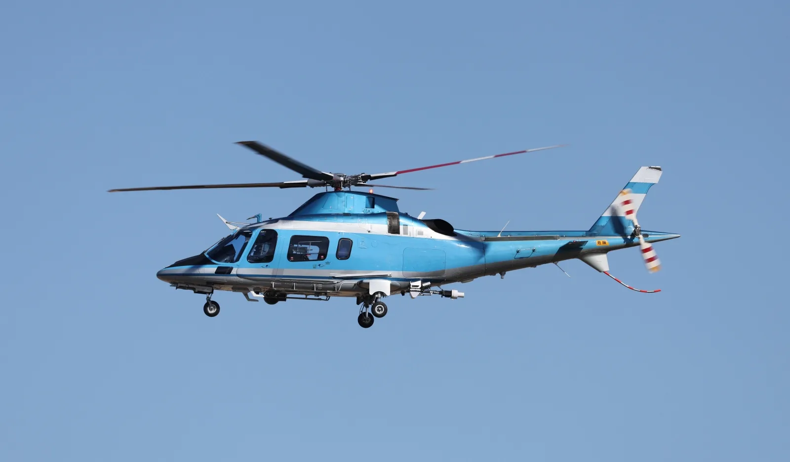 A light blue and white helicopter flies against a clear blue sky.
