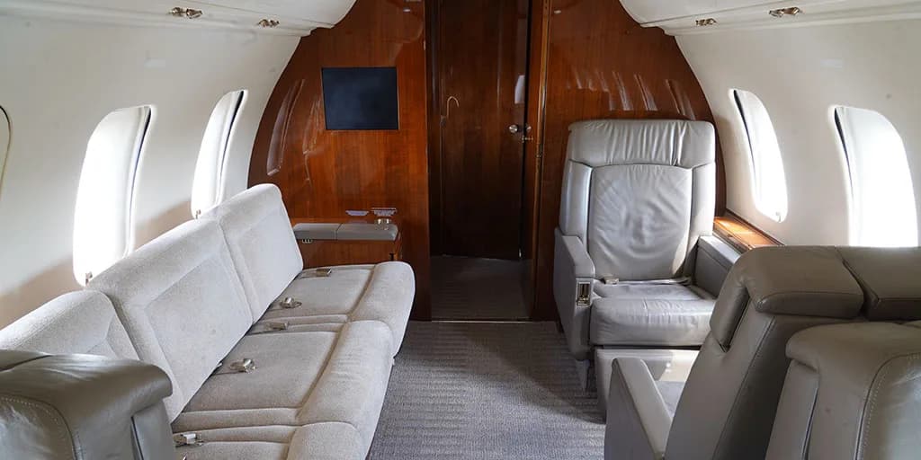 The interior cabin of a private jet features a light grey sofa and a single leather seat, with wooden paneling and a small screen on the rear bulkhead.