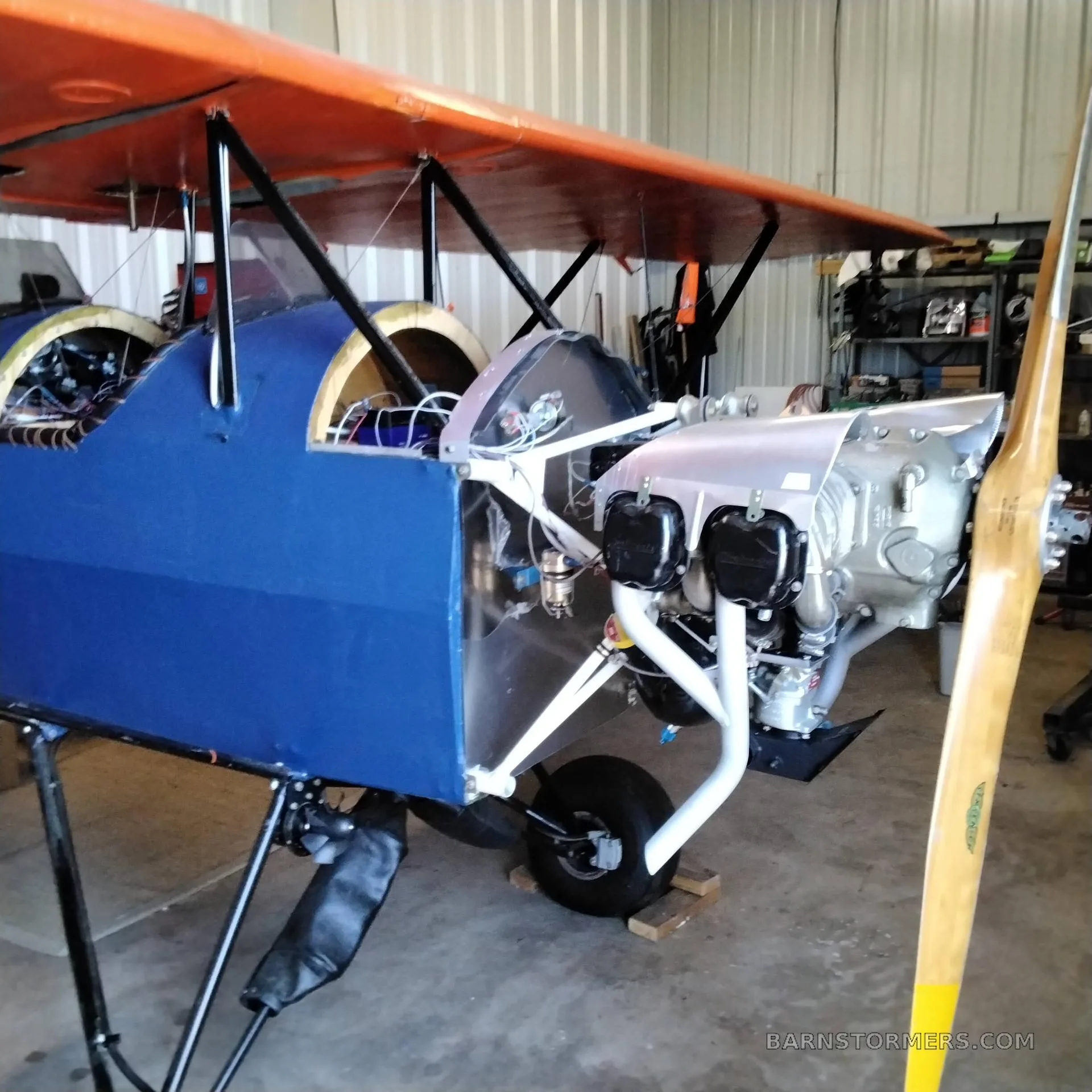 A blue and orange biplane with an exposed engine and wooden propeller, undergoing assembly or maintenance in a hangar.