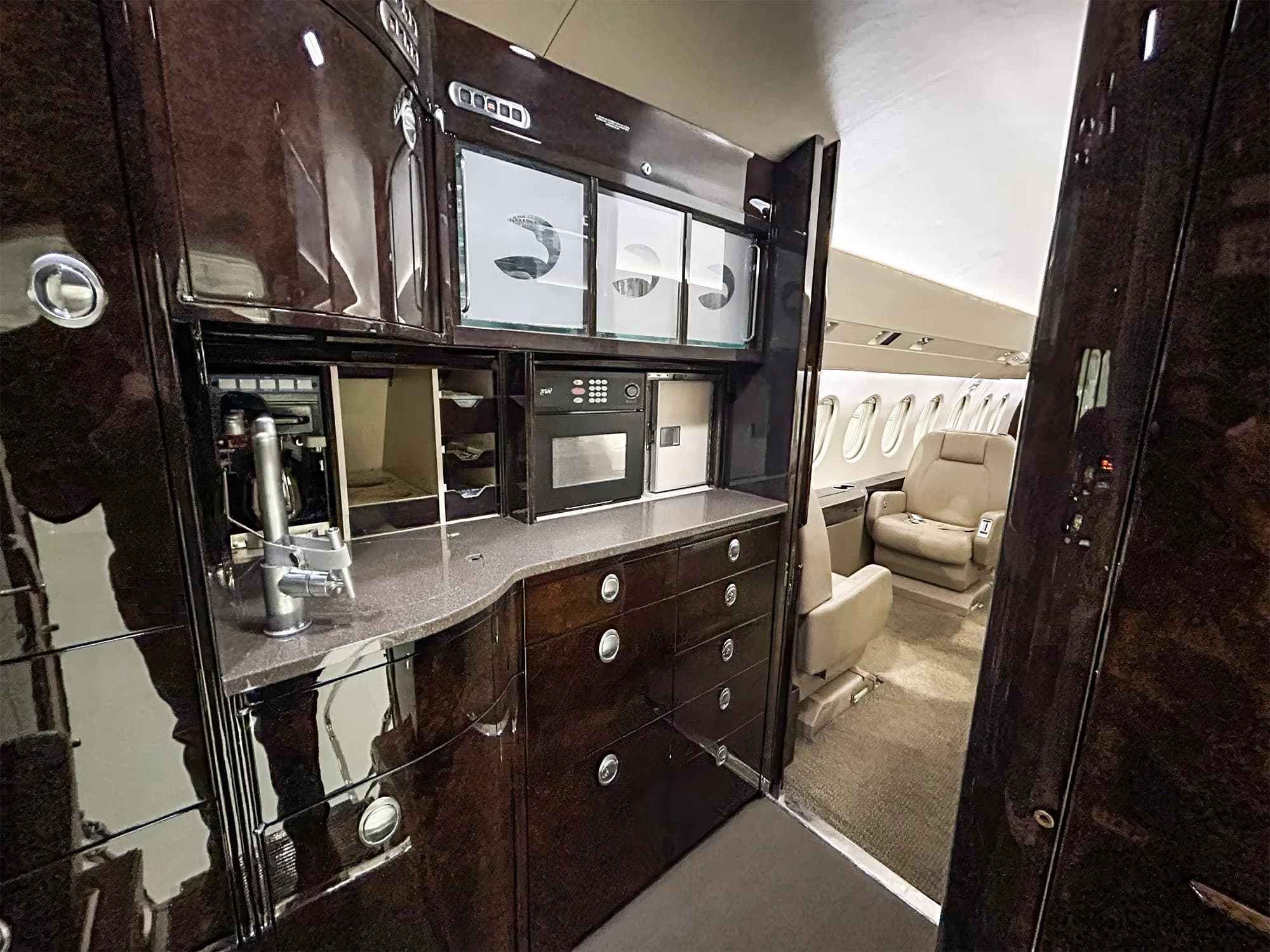A detailed view of the galley in a Dassault Falcon 900EX, featuring dark wood cabinetry, a sink, microwave, and storage compartments.