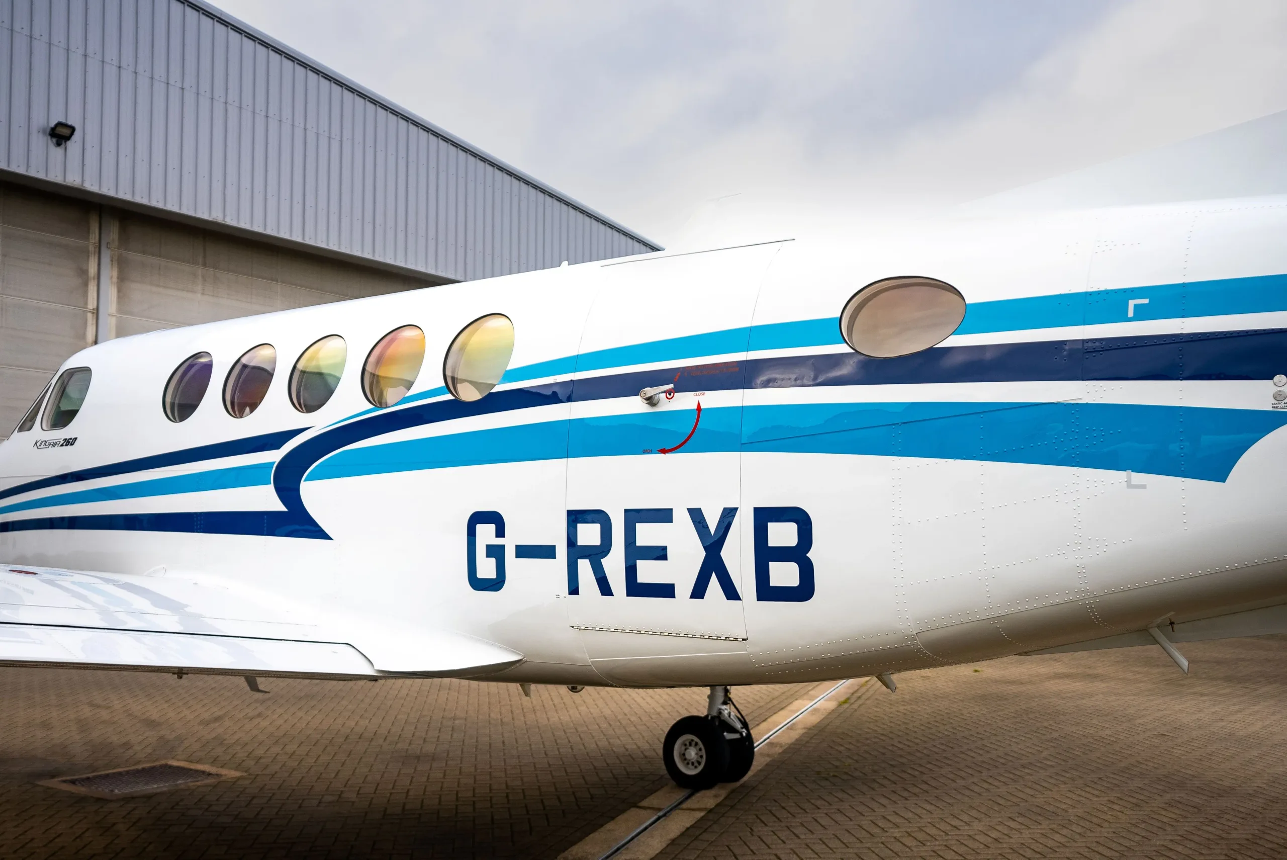 A close-up side view of the white and blue fuselage of a Beechcraft King Air 260, displaying the registration G-REXB.